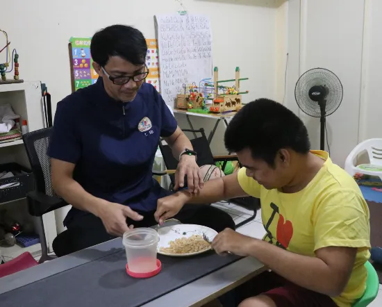 Therapist guiding a young man in practicing independent eating skills with a fork and spoon